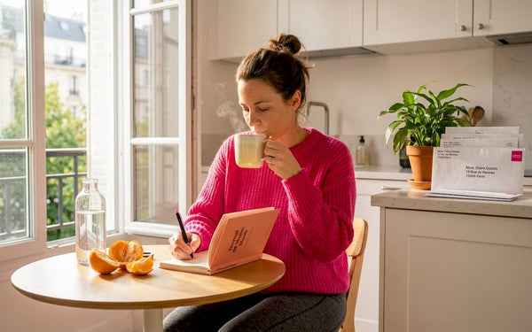 Une femme prend le temps d’écrire dans son carnet, installée à la table de la cuisine, baignée par la lumière du soleil.